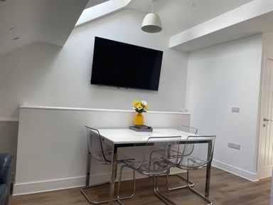 A dining area with a white table and four transparent chairs a wall-mounted television and a yellow vase with flowers on the table at Golygfa'r Gastell Castle View in Criccieth