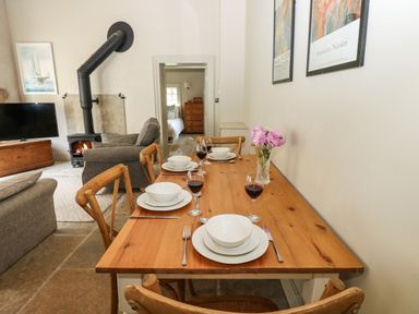 A wooden dining table with plates wine glasses and a vase of flowers near a living room with a wood stove and a doorway to a bedroom at The Courtyard in Kirklinton near Longtown