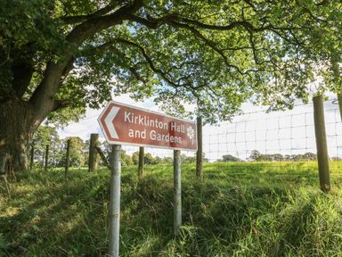A signpost under a tree pointing to Kirklinton Hall and Gardens with a wire fence and grassy field