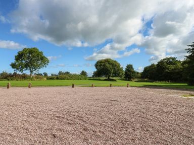 A gravel area with wooden posts separating it from a grassy field with trees under a partly cloudy sky at The Courtyard in Kirklinton near Longtown