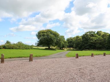 A gravel parking area with wooden posts leading to a narrow road with green fields and trees at The Courtyard in Kirklinton near Longtown