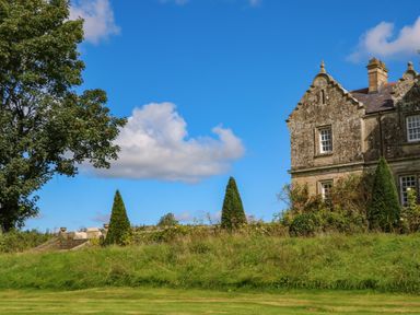 A stone building with a lawn trees and bushes under a blue sky at The Pavillion in Kirklinton near Longtown