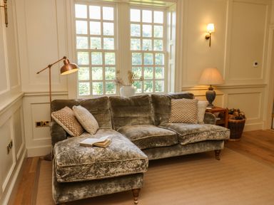 A living room with a velvet sofa with cushions and a book a floor lamp a table lamp a basket of firewood and windows at The Pavillion in Kirklinton near Longtown