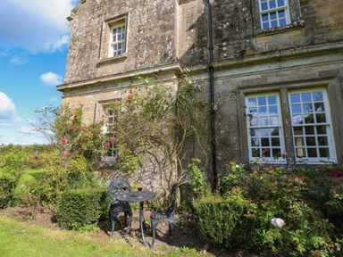 An outdoor seating area with a small table and two chairs beside a stone house with windows and climbing roses at The Pavillion in Kirklinton near Longtown