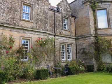 A stone building exterior with white framed windows climbing roses and a metal table with two chairs on grass at The Pavillion in Kirklinton near Longtown