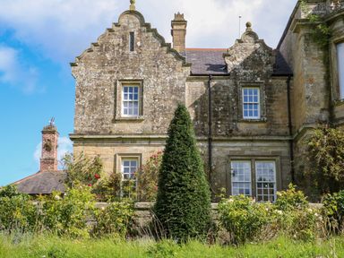 The exterior of a stone house with multiple windows a chimney and a garden with bushes and a tall conical tree at The Pavillion in Kirklinton near Longtown