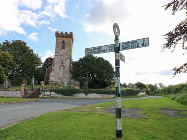 A road signpost on a grassy area near a church with a tower and gravestones at The Pavillion in Kirklinton near Longtown