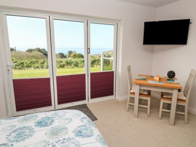 A room with a bed near sliding glass doors showing an outdoor view and a small dining table with two chairs and a wall-mounted television at Greenside Studio in Praa Sands
