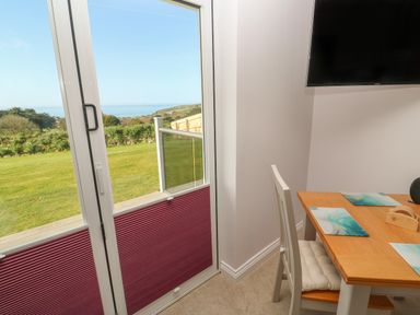 A dining area with a wooden table and white chair near a glass door showing a grassy garden and distant hills at Greenside Studio in Praa Sands