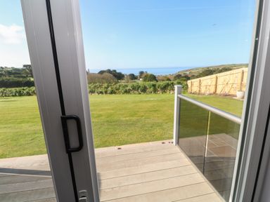 A view through glass door onto a wooden patio overlooking a grass lawn and hills with the sea in the distance at Greenside Studio in Praa Sands
