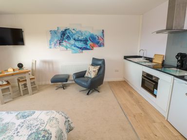 A room with a bed a dining table with two chairs a blue armchair with a matching footstool a kitchen area with a sink oven and refrigerator and a wall-mounted television at Greenside Studio in Praa Sands