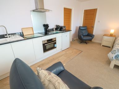 A compact kitchen area with an oven, sink, and countertop appliances next to two blue chairs and a bedside table with a lamp at Greenside Studio in Praa Sands