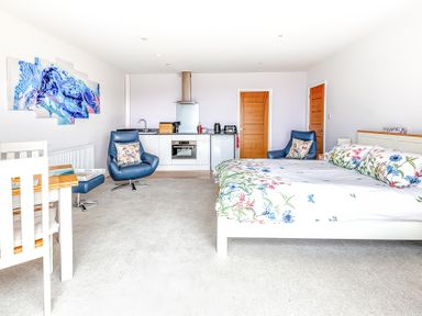 A bedroom with a double bed floral bedding two blue chairs a small kitchen unit and a dining table at Greenside Studio in Praa Sands