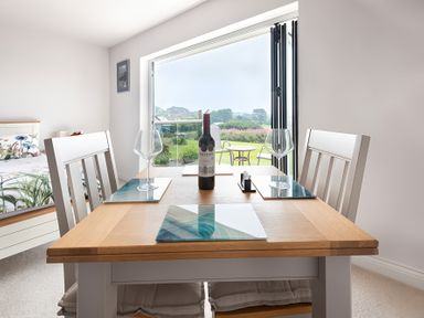 A dining table with two chairs wine bottle and glasses by a window overlooking a garden at Greenside Studio in Praa Sands