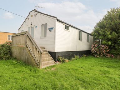 A white mobile home with steps leading to a door on a grassy lawn with bushes at Suzannes Beach Hut in Bacton near Walcott
