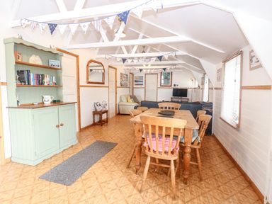 A dining area with wooden table and chairs a green bookshelf and seating area with sofas and a television at Suzanne's Beach Hut in Bacton near Walcott