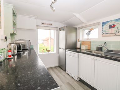 A kitchen with black countertops white cabinets a refrigerator dishwasher sink and small windows at Suzanne's Beach Hut in Bacton near Walcott