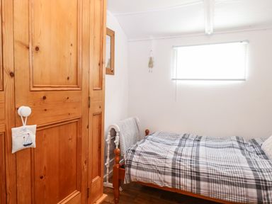 A bedroom with a wooden wardrobe a bed with checkered bedding a window with blinds and a wall mirror at Suzanne's Beach Hut in Bacton near Walcott