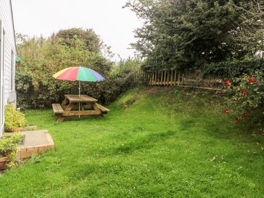 A backyard with a wooden picnic table and multicolored umbrella near a white house with green grass and bushes at Suzannes Beach Hut in Bacton near Walcott