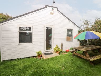 A white exterior wall with a glass door and two windows next to a wooden picnic table with a colorful umbrella on grass at Suzanne's Beach Hut in Bacton near Walcott