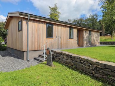 A single-story wooden house with black-framed windows a small staircase and a stone wall in a grassy yard at Millstream Ridge in Haverthwaite