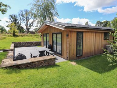 A wooden cabin with sliding glass doors and a stone patio with a table and benches at Millstream Ridge in Haverthwaite