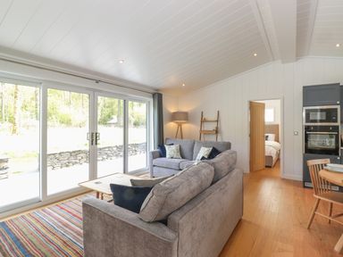 A living room with two grey sofas a striped rug a wooden coffee table large windows and an open door to a bedroom at Hawthorn Croft in Haverthwaite