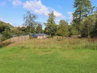 A grassy field with trees and wooden buildings with solar panels behind a wooden fence at Hawthorn Croft in Haverthwaite