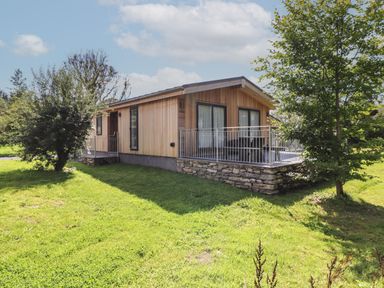 A wooden house with a stone base and metal railing around the porch in a grassy yard with trees at Ash Thwaite in Haverthwaite