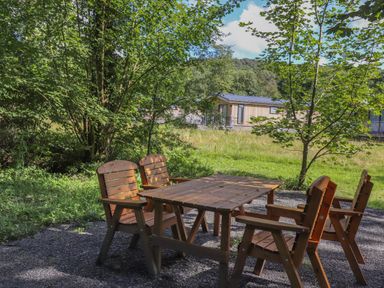 An outdoor wooden table with four wooden chairs on a gravel patch surrounded by trees and grass at Ash Thwaite in Haverthwaite
