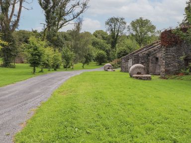 A gravel path beside a green lawn with small trees and stone buildings with millstones at Ash Thwaite in Haverthwaite