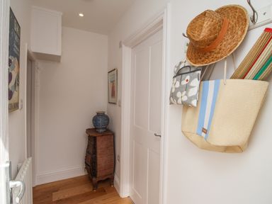 A hallway with a chest of drawers and a blue vase a closed white door and hats and bags hanging on wall hooks at The Corner House in Weymouth