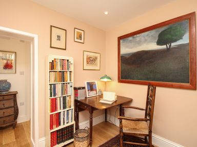 A small study area with a wooden desk and chair beside a bookshelf filled with books and framed pictures on the walls at The Corner House in Weymouth