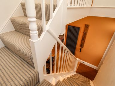 A staircase with striped carpet and white railing overlooking an orange wall with a black radiator and wooden coat hooks at The Corner House in Weymouth