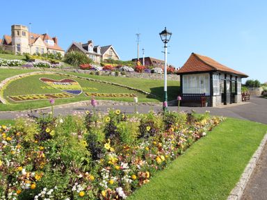A landscaped garden with flowers a paved path a small building and houses in the background at The Corner House in Weymouth