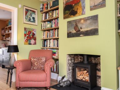 A corner with a red striped armchair a side table with a lamp a wood stove with logs and paintings on the green walls at The Corner House in Weymouth