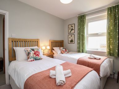 A bedroom with two single beds with floral pillows and orange throws a window with green curtains and two bird pictures on the wall at The Corner House Weymouth