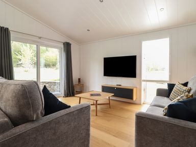 A living room with two grey sofas a wooden coffee table a wall-mounted television and large windows with grey curtains at The Brambles in Haverthwaite