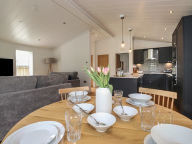 A dining table set with dishes and glasses a vase with flowers a grey sofa and a kitchen area with dark cabinets at The Brambles in Haverthwaite