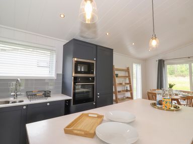 A kitchen with dark cabinets a built in oven and microwave a white countertop with plates and a wooden tray and a dining area with wooden chairs and table at The Brambles in Haverthwaite
