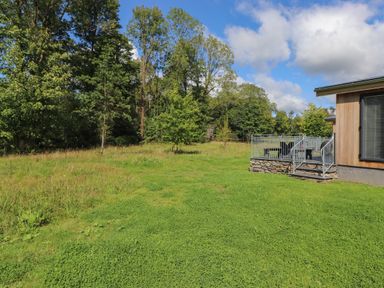 A grassy backyard with trees and a deck attached to a wooden house at The Brambles in Haverthwaite