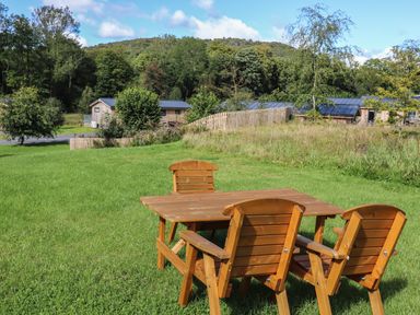 An outdoor area with wooden table and chairs on grass near trees and houses at The Brambles in Haverthwaite