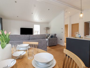 A dining table with dishes glasses and a vase with flowers in an open living space with a gray sofa and a television at The Burrow in Haverthwaite