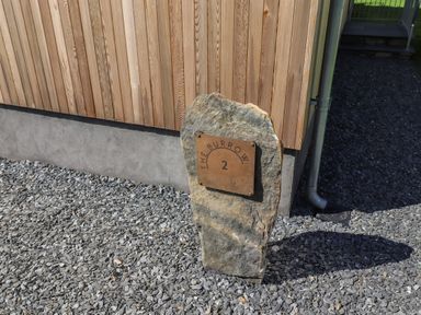A stone with a metal plaque showing The Burrow 2 outside a building with wooden siding and gravel ground at The Burrow in Haverthwaite