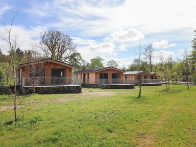 Three wooden cabins with stone foundations and metal railings surrounded by grass and trees at Blackthorn Copse in Haverthwaite