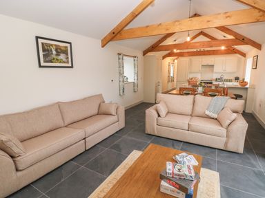 A living room with two beige sofas a wooden coffee table with board games and an open kitchen with wooden beams at The Byre in St Brides Major