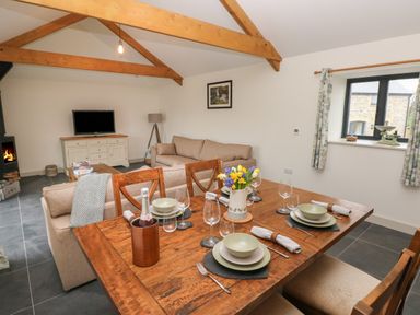 A dining table set with plates glasses and napkins in an open living and dining room with sofas a television and a fireplace at The Byre in St Brides Major