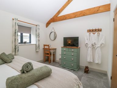 A bedroom with a bed, rolled towels, a green chest of drawers with a TV, a wooden desk with a chair, a round mirror, wall hooks with two white robes, and a window with curtains at The Byre in St Brides Major