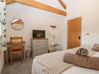 A bedroom with a wooden chair and desk, a round mirror, a dresser with a TV, bathrobes on hooks, and a bed with folded towels at The Byre in St Brides Major