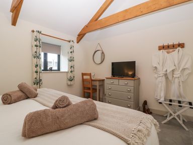A bedroom with a bed towels a window with curtains a desk a chair a mirror a chest of drawers with a television and bathrobes on hooks at The Byre in St Brides Major